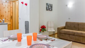 A modern living room with a brown sofa, a dining table with orange cups and patterned plates, a small side table with a vase of red flowers, and a wooden door.