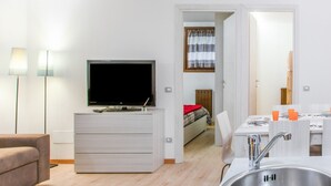 A modern, well-lit room with a flat-screen TV on a wooden cabinet, a sofa, a dining table with a sink, and a view into the adjacent bedroom.
