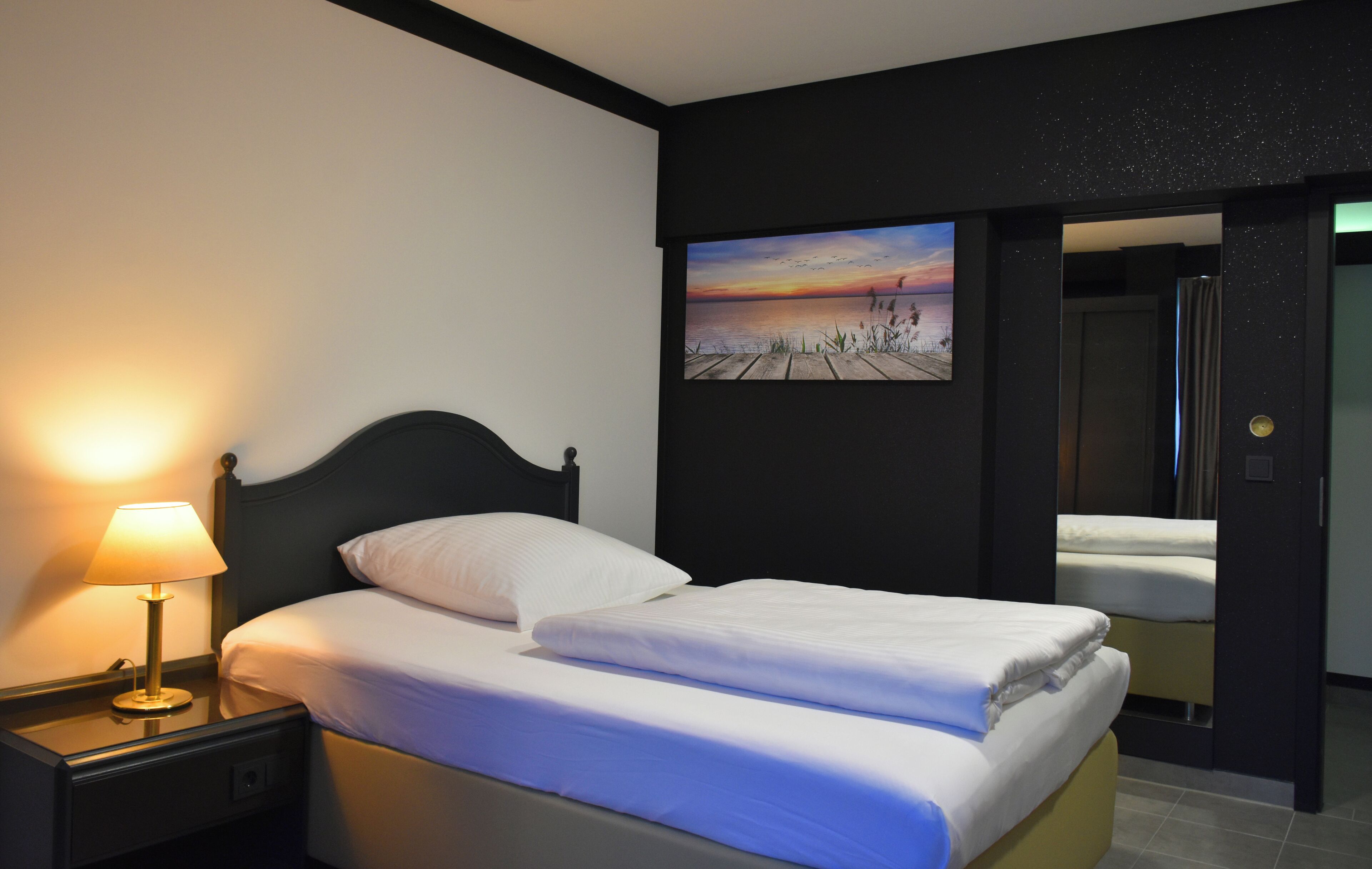 Habitación de hotel con cama, mesita de noche con lámpara, pintura de un atardecer y zona de ducha.