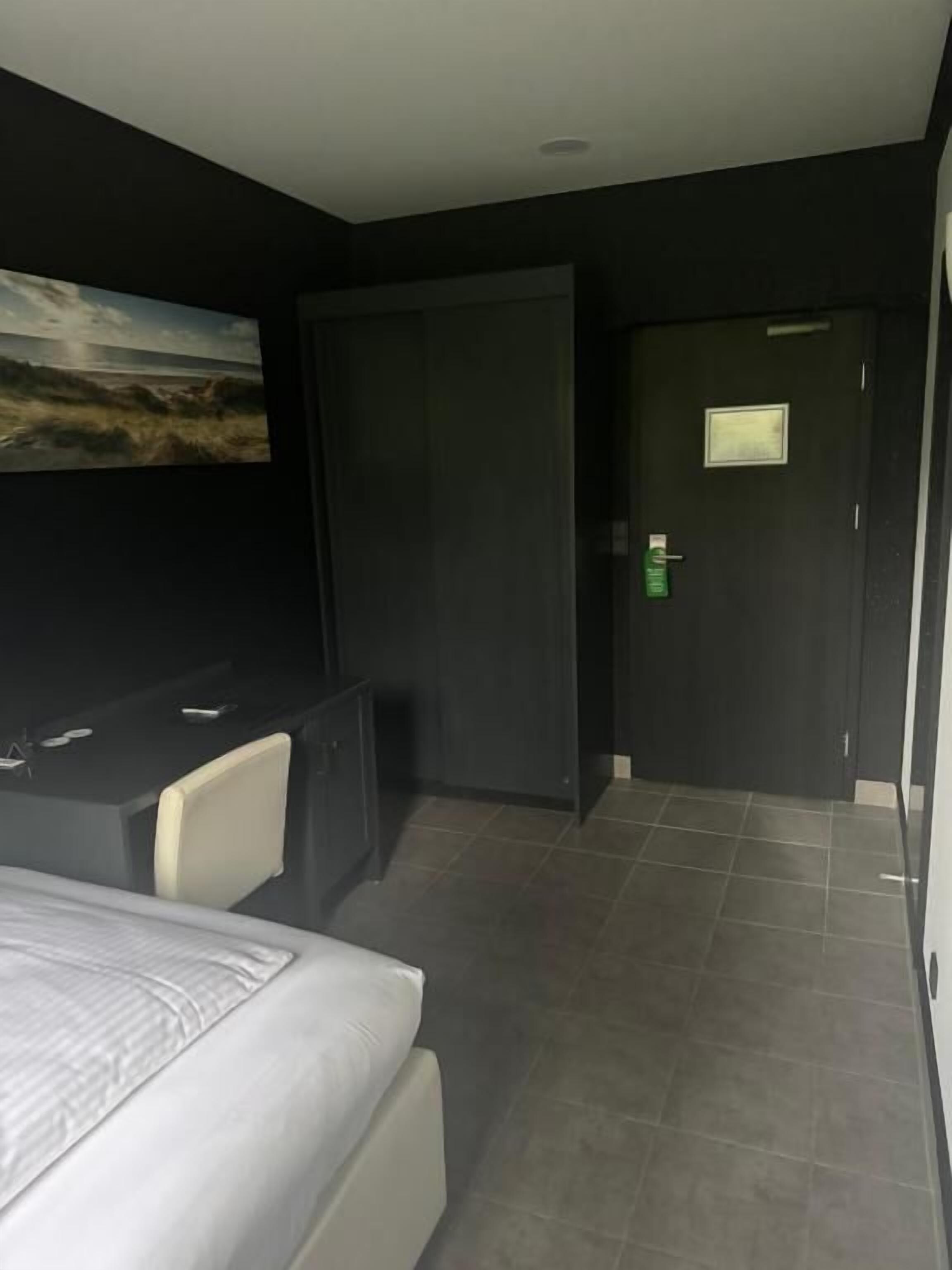 A modern hotel room with a bed, a desk, and a chair. There is a painting on the wall and a door with a light switch.