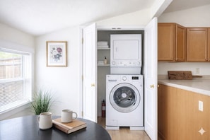 Eine kleine Küche mit Waschmaschine, Holzschränken, einem gerahmten Bild an der Wand und einem Tisch mit Tasse und Buch.