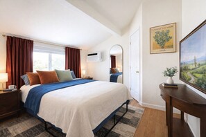 Ein Hotelzimmer mit Bett, Nachttisch mit Lampe, Spiegel, Gemälde und Fenster mit Vorhängen.