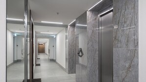 A modern hallway with marble walls, a glass elevator, and a long corridor with doors at the end.