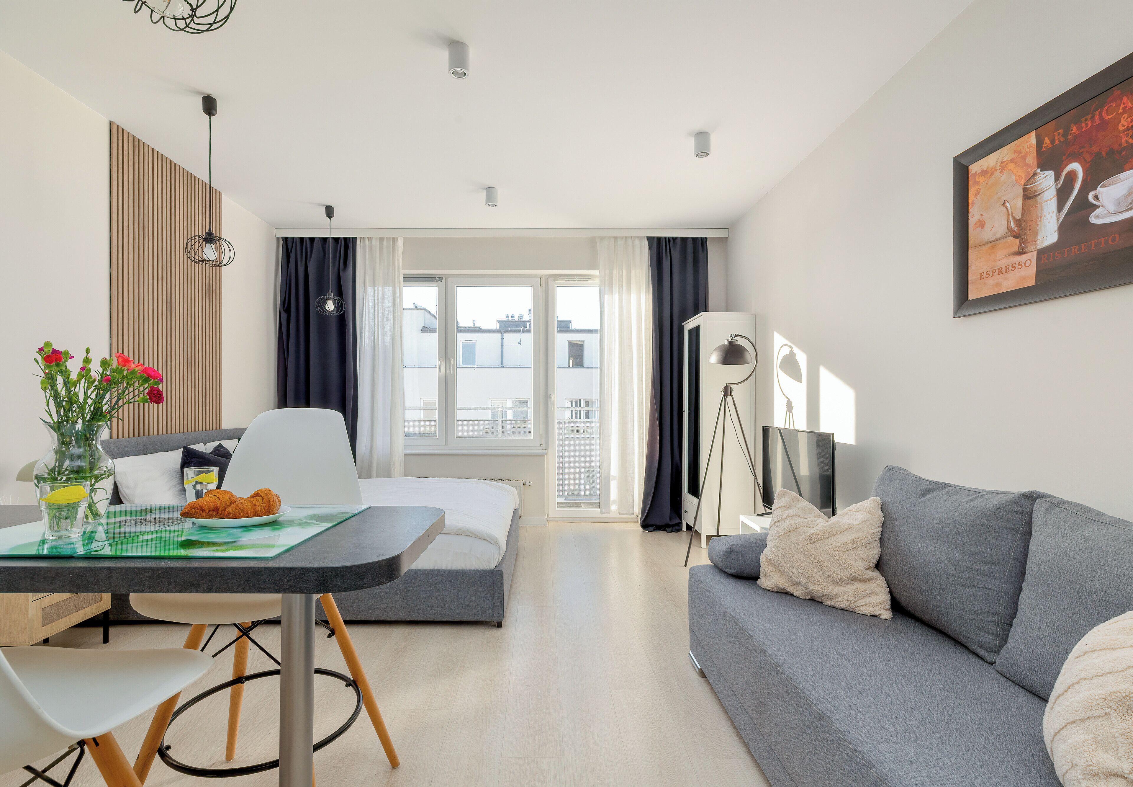 A modern living room with a grey sofa, a dining table with a green placemat, a white chair, and a large window with curtains.