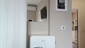 Pieni, valkoinen jääkaappi käytävällä, jonka seinä on beigen värinen. Seinällä on myös kiinnitetty televisio ja kehystetty taulu.