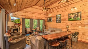 A cozy cabin interior with a fireplace, leather sofas, a wooden dining table, and a large window overlooking greenery. - Cliffside Mountain View - Private indoor pool, gorgeous views, theater, game room, and more! (Sevierville)