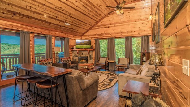 A cozy cabin interior with a wooden interior, a fireplace, a dining area, and large windows offering a view of the outdoors. - Cliffside Mountain View - Private indoor pool, gorgeous views, theater, game room, and more! (Sevierville)