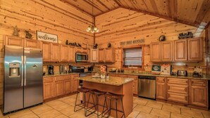 A spacious kitchen with wooden cabinets, a stainless steel refrigerator, a microwave, and a sink. There are two bar stools at the island, and a sign that reads "Welcome" above the refrigerator. - Cliffside Mountain View - Private indoor pool, gorgeous views, theater, game room, and more! (Sevierville)