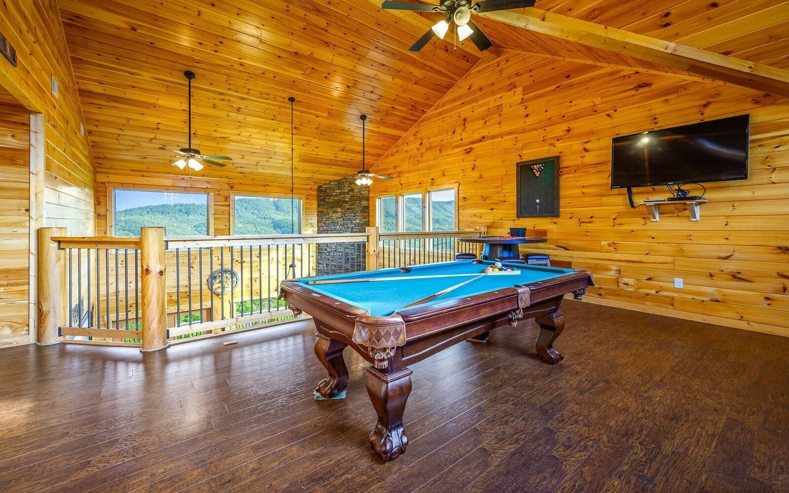 A wooden interior with a pool table, a balcony with a railing, and a television mounted on the wall.