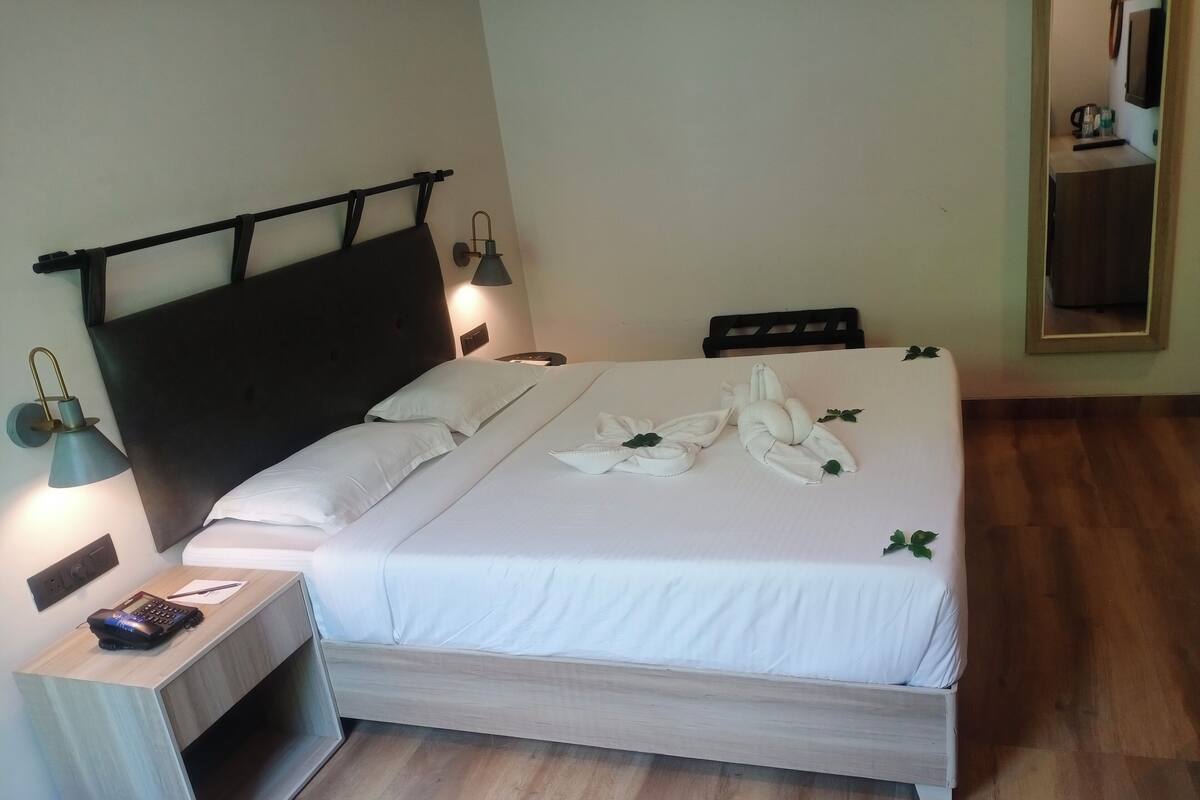 Una cama bien tendida con sábanas y toallas blancas dispuestas en un diseño decorativo, una mesita de noche con una lámpara y un suelo de madera.