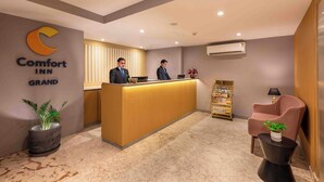 Uma área de recepção de hotel, com balcão, dois funcionários, uma planta, uma estante de livros e uma poltrona.