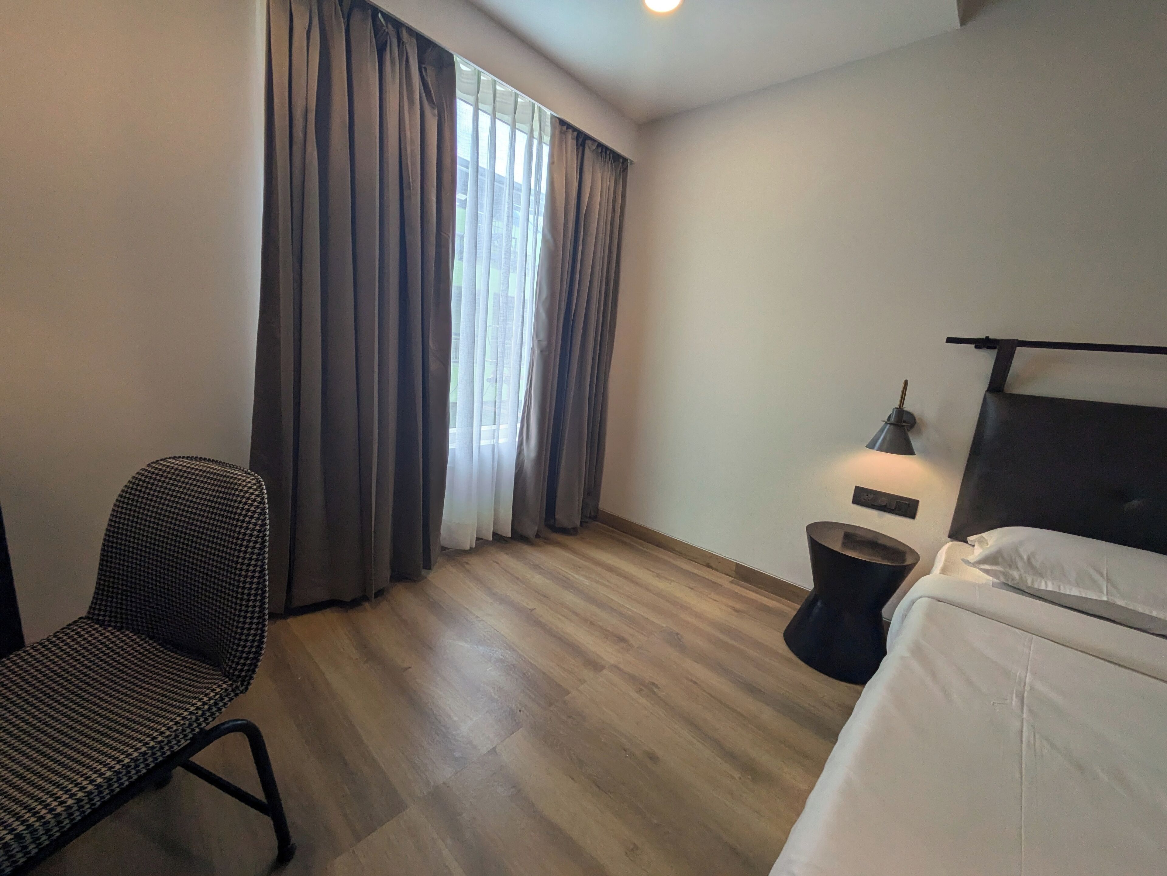 A hotel room with a bed, a chair, a bedside table with a lamp, a window with curtains, and a wooden floor.