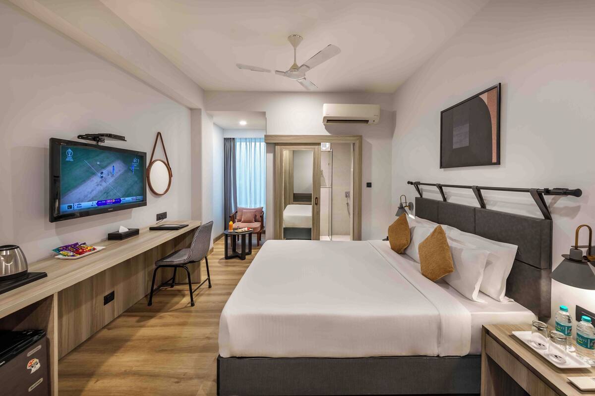 Habitación de hotel con cama, mesita de noche, lámpara, televisor, escritorio, silla y ventilador de techo.