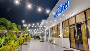 Een hotelentree met een bord waarop "THE MANSION" staat, omgeven door bloempotten en lichtslingers.