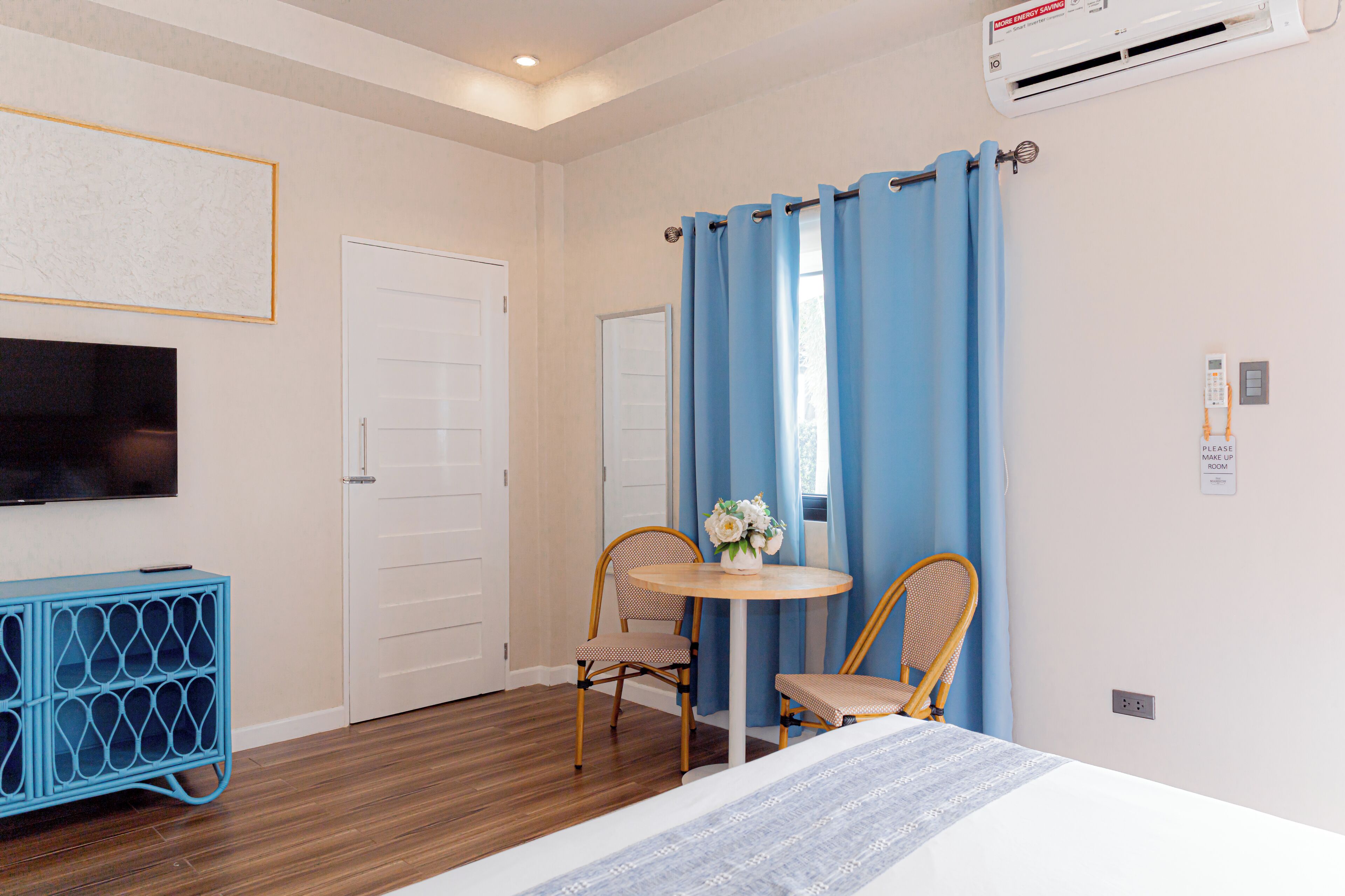 A hotel room with a bed, a TV, a blue wall unit, two chairs, a table with a vase, and a window with blue curtains.