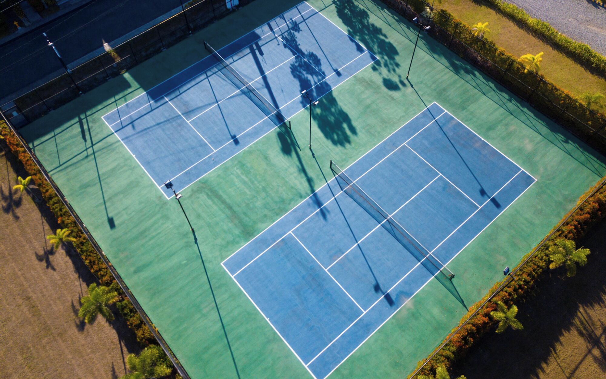 An aerial view of a tennis court with multiple courts, surrounded by greenery and a fence.