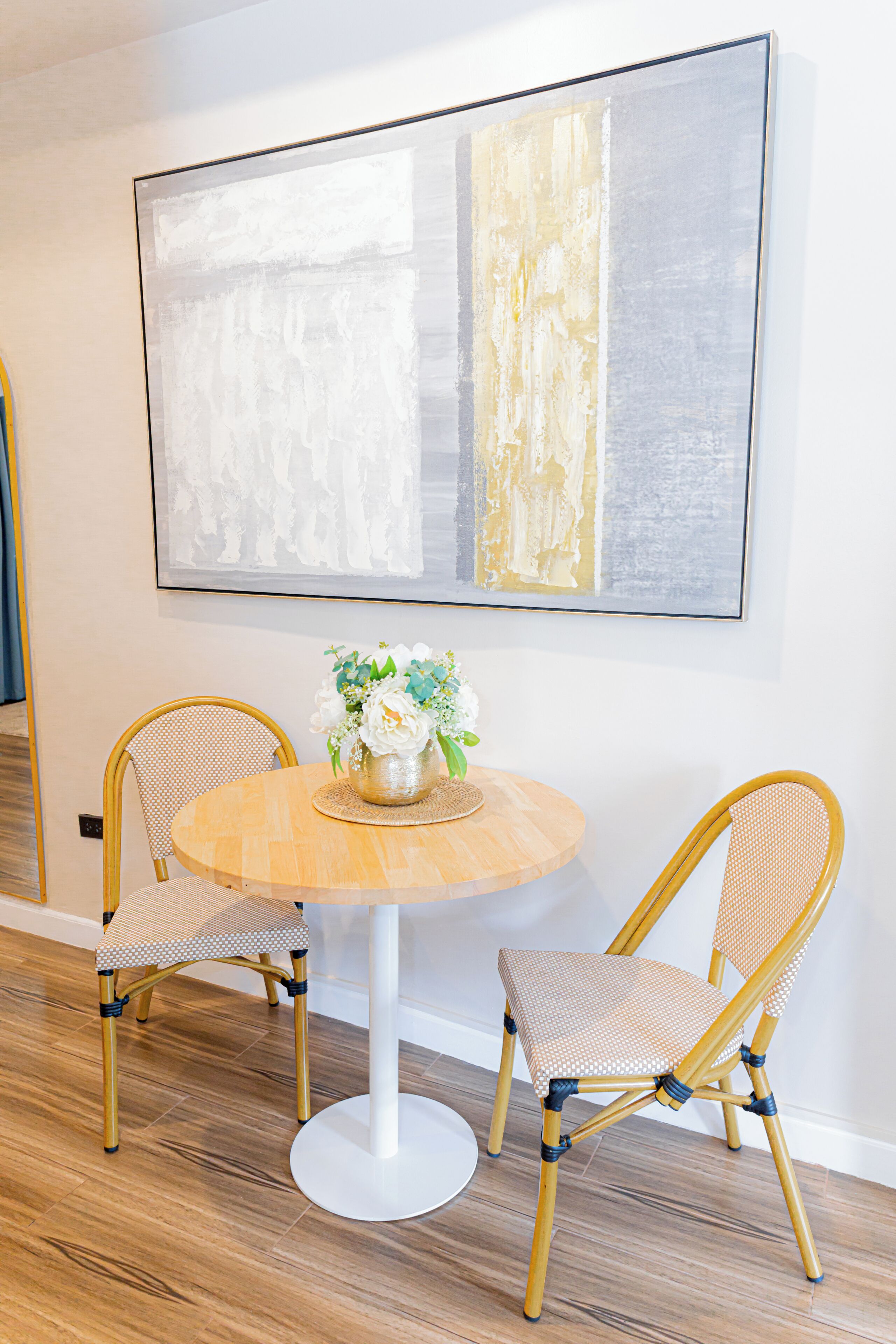 A dining area with a round table, two wicker chairs, and a large abstract painting on the wall.