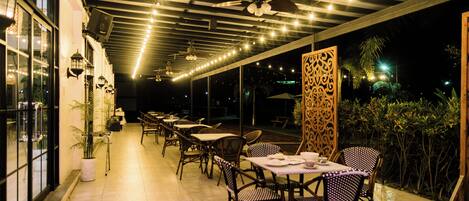 Un espacio de comedor al aire libre con mesas y sillas, ventiladores de techo y luces colgantes.
