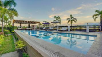 A swimming pool with lounge chairs and umbrellas, surrounded by palm trees and a pergola.