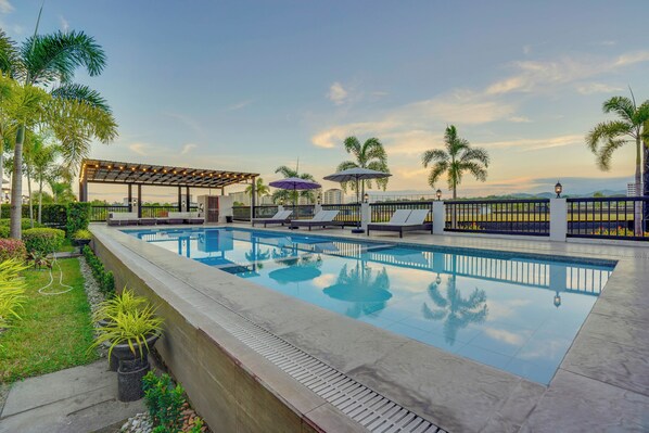 A swimming pool with lounge chairs and umbrellas, surrounded by palm trees and a pergola.