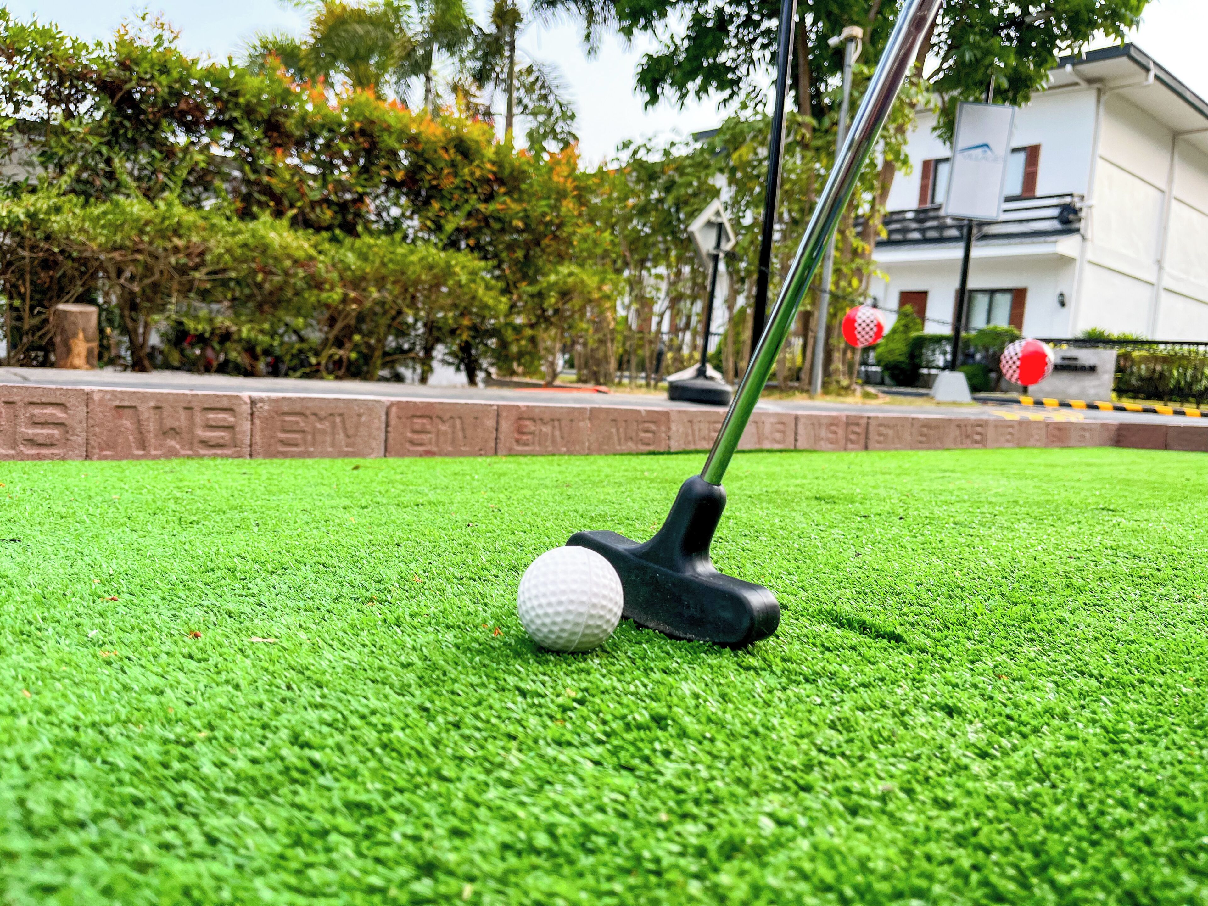 A golf ball on artificial grass with a golf club nearby, a stone wall with inscriptions, and a building in the background.