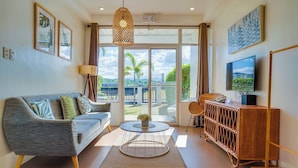Een moderne woonkamer met een grijze bank, een ronde salontafel, een rieten bijzettafeltje, een televisie, en een balkon met uitzicht op palmbomen en een brug.