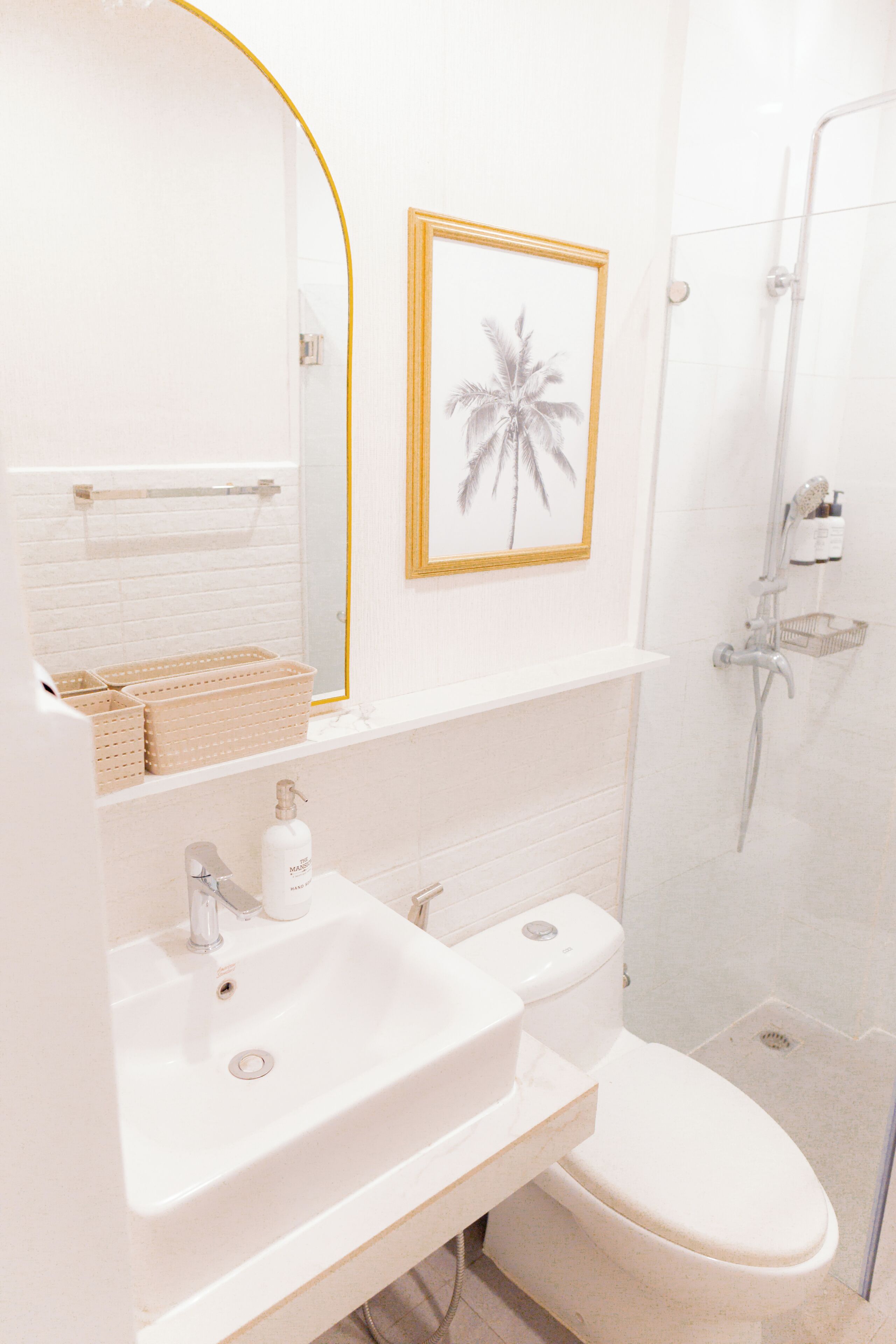 A bathroom with a white sink, toilet, and shower. There is a framed picture of a palm tree on the wall.