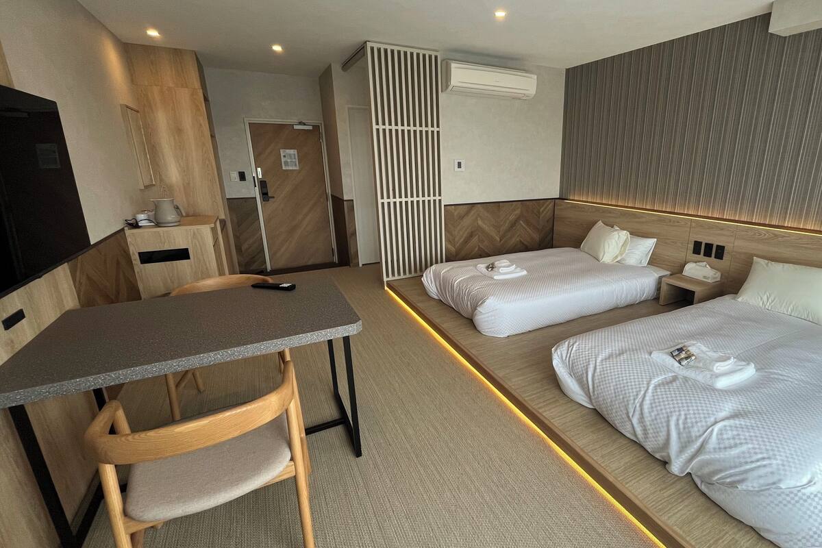 A hotel room with two beds, a desk, a chair, an air conditioning unit, and a TV.