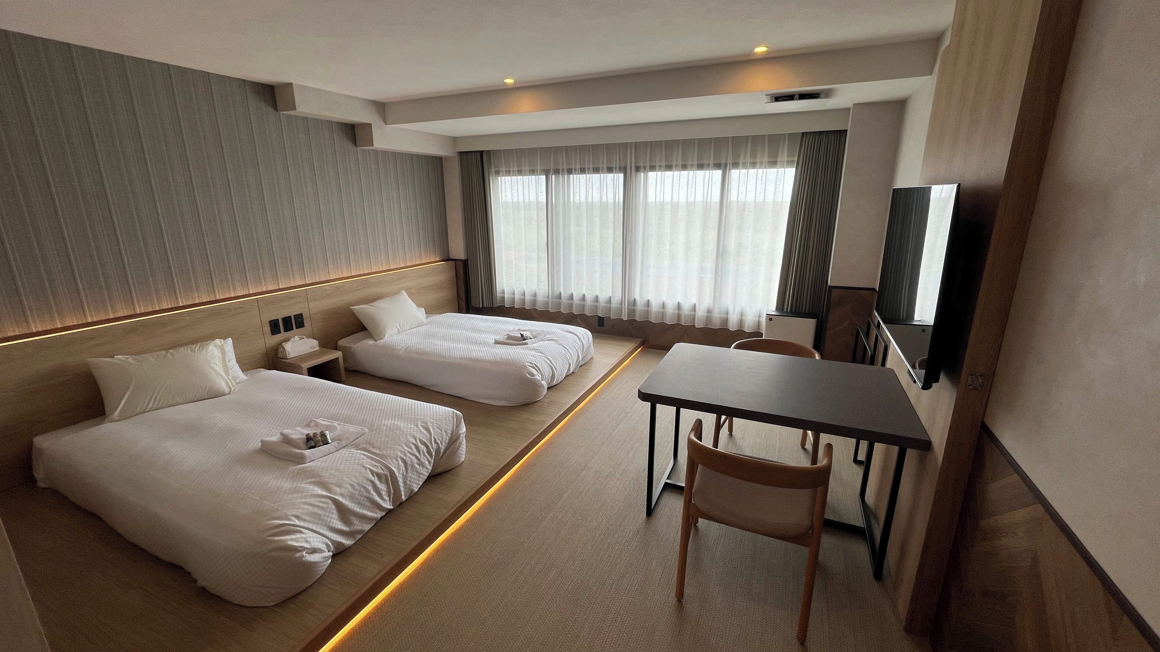 Habitación de hotel con dos camas, un escritorio, una silla, un televisor y un ventanal grande con cortinas.