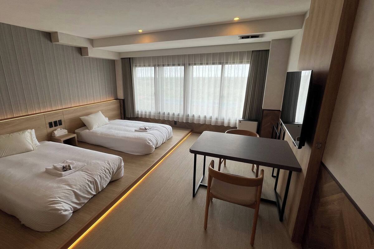 A hotel room with two beds, a desk, a chair, a TV, and a large window with curtains.