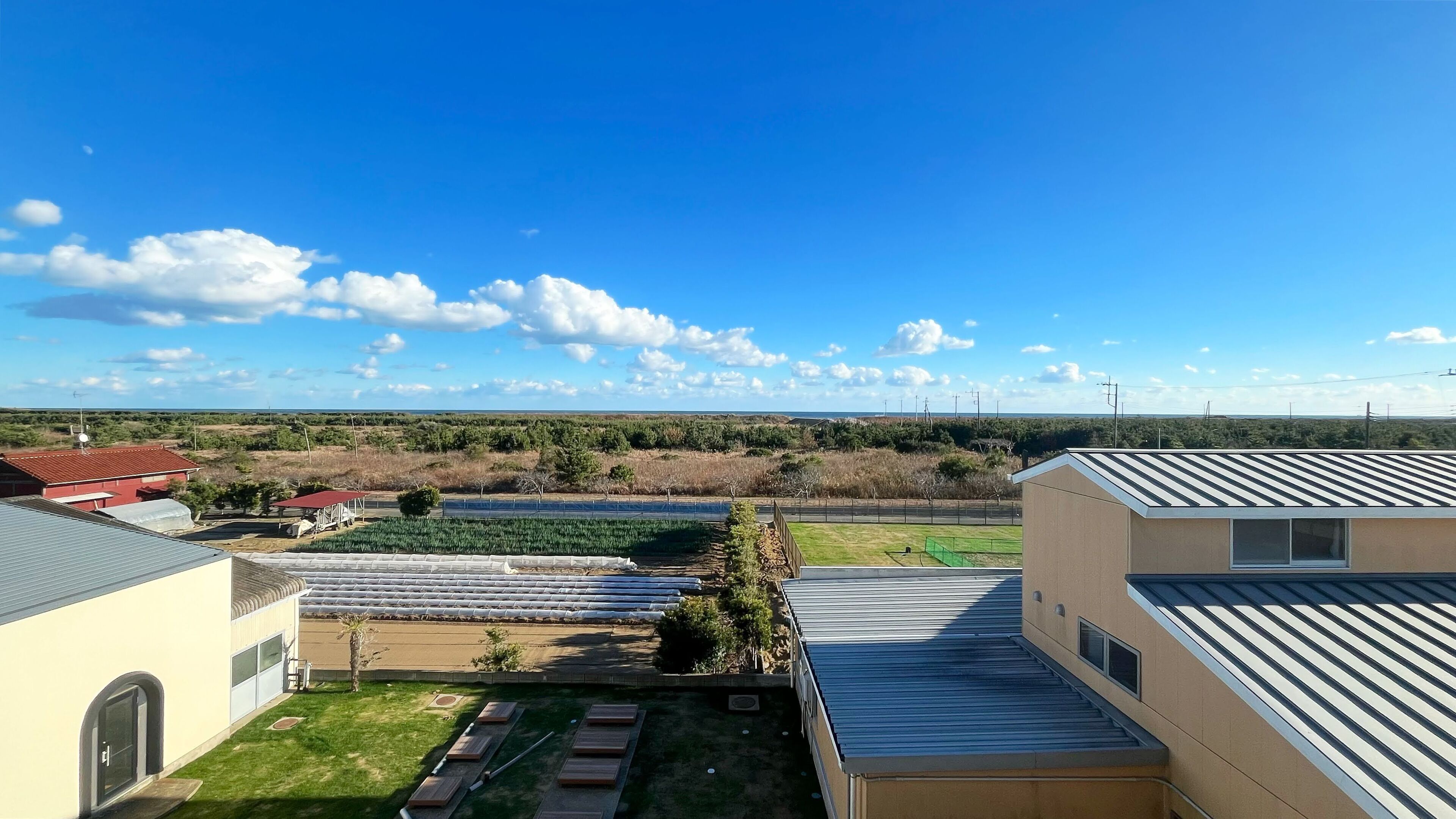 A residential area with a view of a green field and distant trees. There are houses with different roof styles and a clear sky.