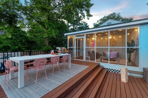 An outdoor deck with a white table and pink chairs, a wooden bench, and a glass-enclosed room with a round wooden wall art piece.