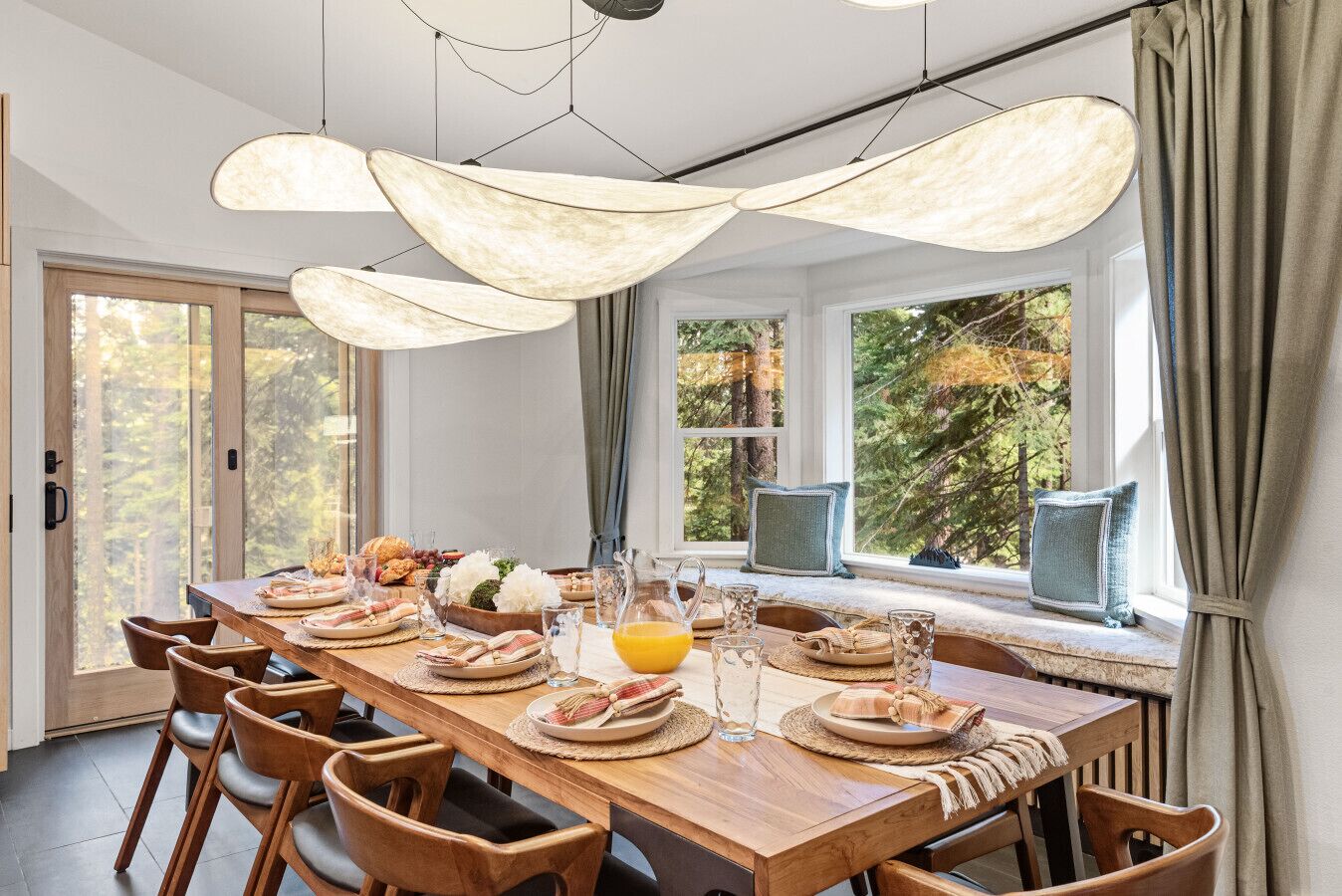 A dining area with a long wooden table set for breakfast, large windows with a forest view, and pendant lights.