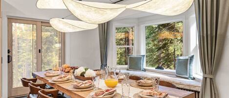 A dining area with a long wooden table set for breakfast, large windows with a forest view, and pendant lights.