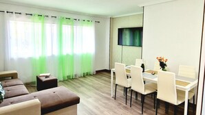 A modern living room with a sofa, a dining table with chairs, a flat-screen TV mounted on the wall, and a large window with sheer curtains. - Los Cristianos, 3 Bedrooms & WiFi Free (LOS CRISTIANOS)