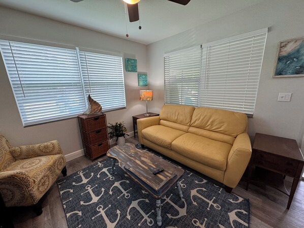 A living room with a sofa, armchair, and a coffee table. There are two windows with blinds, a lamp, and a painting on the wall.