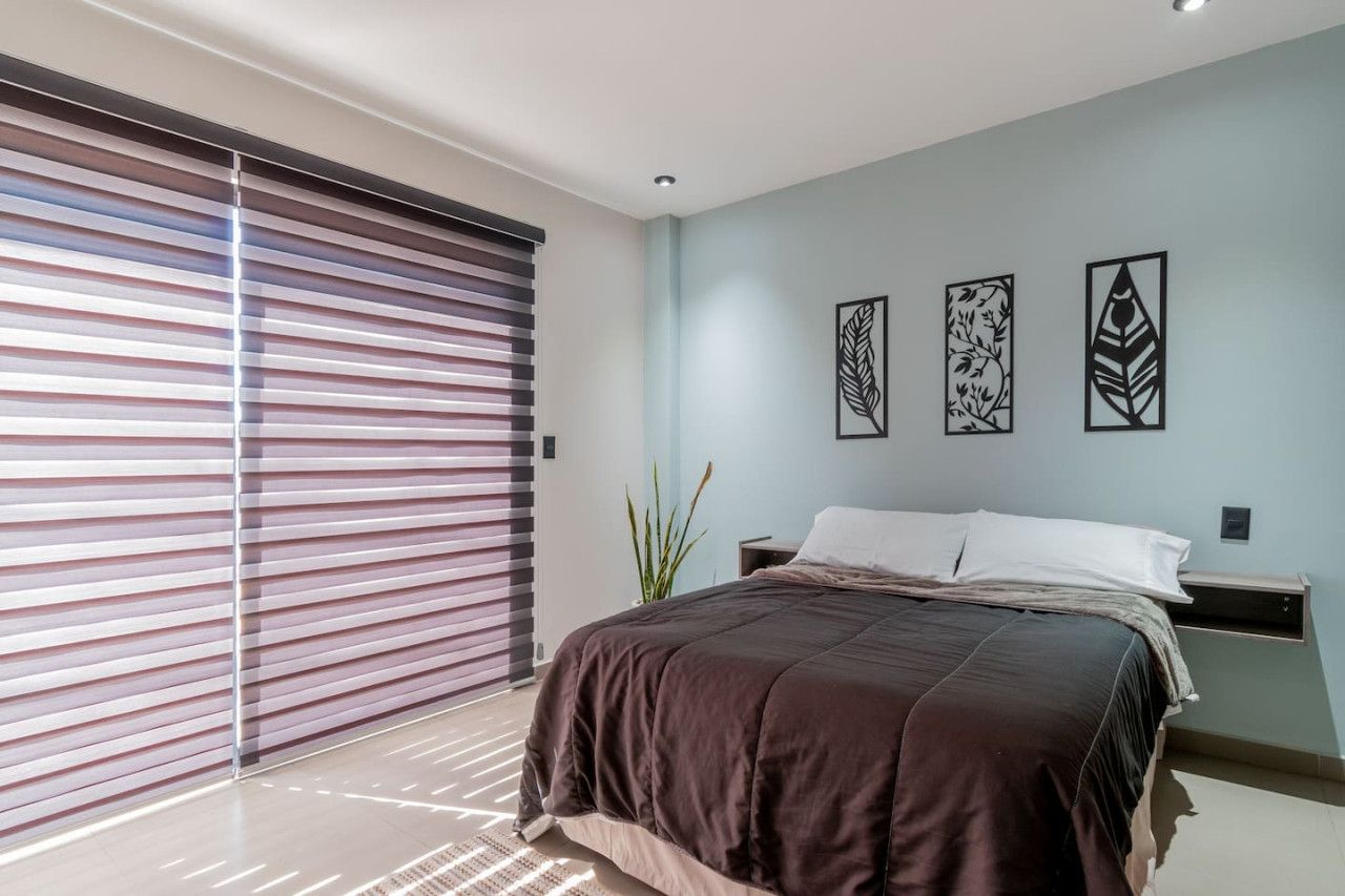 A bedroom with a bed, a nightstand, a wall-mounted shelf, and a large window with blinds.