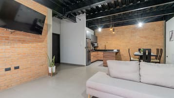 A modern living space with a sofa, a flat-screen TV mounted on a brick wall, a kitchen area with a countertop, and a dining table with chairs.