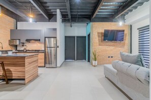 Cozinha moderna com ilha integrada, eletrodomésticos de aço inoxidável e sofá cinza numa sala de estar.