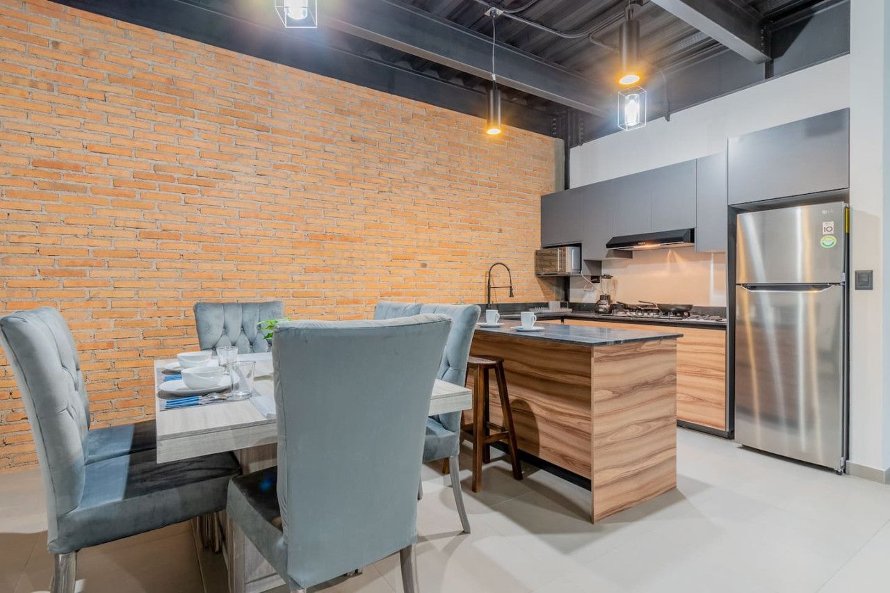 A modern kitchen with a brick wall, stainless steel appliances, and a dining area with grey upholstered chairs.
