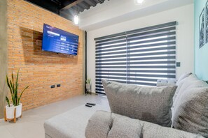 A modern living room with a brick wall, a mounted TV, a large window with blinds, and a grey sofa. - Depa Industrial Moderno y lujoso en Excelente ubicacion 2 (Durango)
