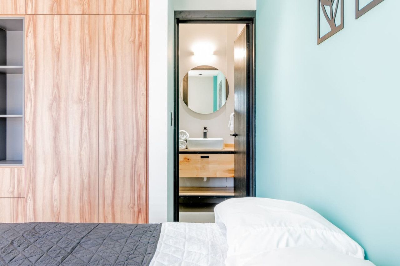 A bedroom with a bed, a wooden wardrobe, a bathroom with a mirror, sink, and towel rack.