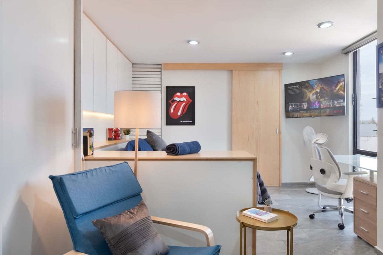 A modern hotel room with a blue armchair, a small round table with a book, a desk with a lamp, and a large poster on the wall.