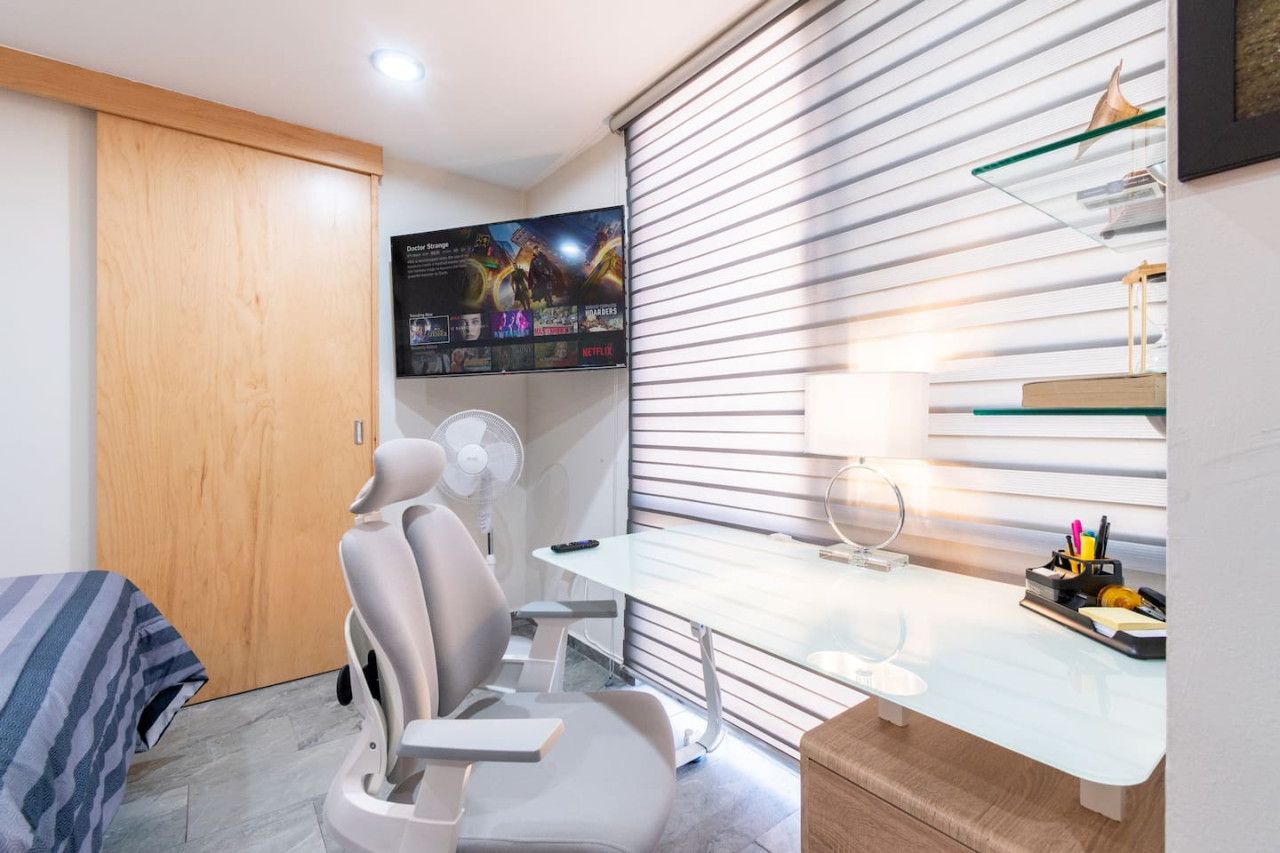 A modern room with a bed, a desk, a chair, a fan, and a wall-mounted TV displaying movie posters.