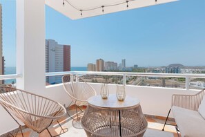 A balcony with wicker furniture, a round table, and a view of city buildings and the sea.