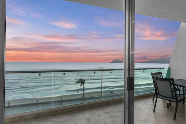 A balcony with a glass railing overlooking a beach at sunset.