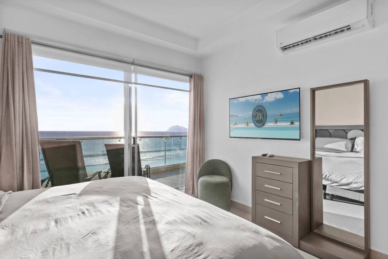 A hotel room with a bed, a chair, a dresser, and a large window offering a view of the ocean.