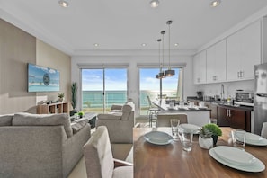 Un salon moderne avec un canapé et une table à manger pour quatre personnes, une cuisine avec des meubles blancs et une grande fenêtre donnant sur l’océan.