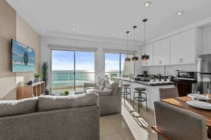 Una cocina moderna con armarios blancos y electrodomésticos de acero inoxidable, un comedor con mesa de madera y taburetes de bar, y una sala de estar con sofá y vista al océano.