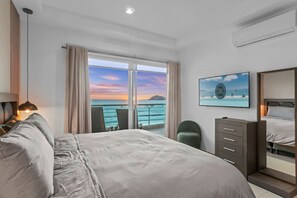 Quarto de hotel com cama, escrivaninha, cadeira, cômoda e uma grande janela com vista para o oceano.